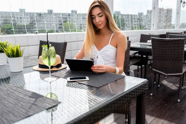 Das Mädchen im Kleid sitzt in einem Sommercafé. Frau hält ein Tablet auf dem Tisch, ein Glas Limettengrün. Video ansehen. Anwendung für soziale Netzwerke. Geschäftsführung per E-Mail. — Stockfoto