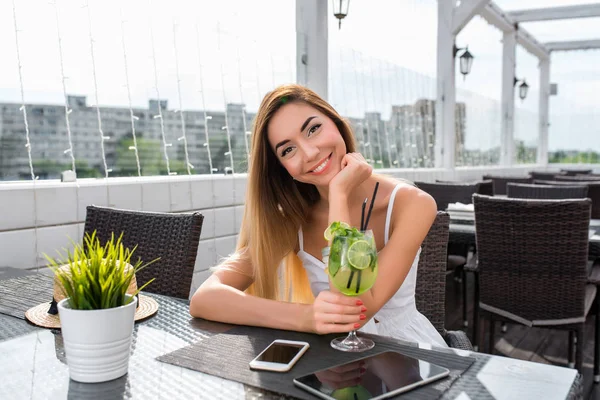 Schöne und glückliche Mädchen lächelt im Sommercafé. Auf dem Tisch liegt ein elektronisches Tablet-Telefon. ein Glas lindgrünes Getränk. posieren, während sie auf Freunde warten. Frühstück nach dem Mittagessen im Freien. — Stockfoto