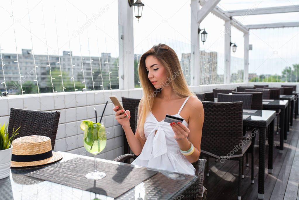 Beautiful girl smiling in summer cafe. In hands of plastic credit card, pay for breakfast and lunch app in Internet. Contactless payment smartphone. Writes pin code from card in open air.