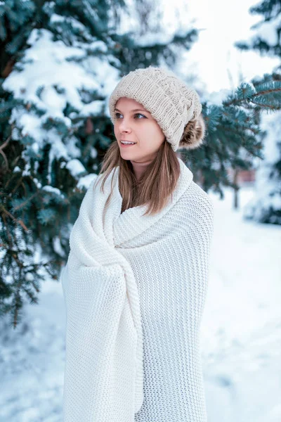 Una bella e giovane ragazza sta in piedi in inverno nella foresta avvolta in un plaid bianco. Cappello caldo, alberi verdi nella neve sullo sfondo. Riposo sulla strada nella sera invernale al resort . — Foto Stock