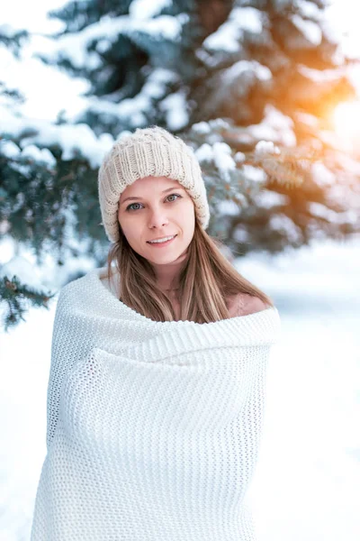 Beautiful happy young girl standing in winter in forest wrapped up white plaid. Warm hat, green trees in snow in background. Rest on street in the winter evening at the resort. Smiles a good mood. — Stock Photo, Image
