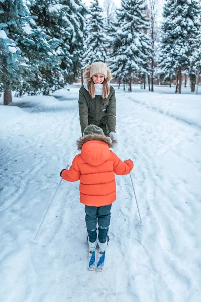 3-5-летний мальчик катается и ходит на игрушечных лыжах с молодой матерью. Зимний лес дрейфует и дрейфует на заднем плане. Первые шаги в спорте активный образ жизни ребенка. Уход и поддержка родителей зимой . — стоковое фото