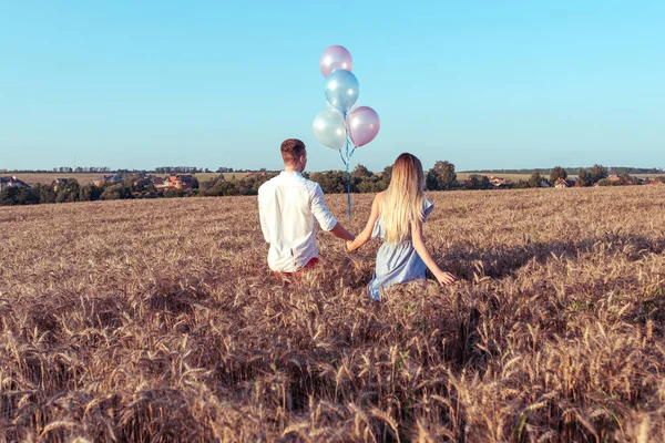 Mulher e homem vão para o campo de trigo de verão. Espaço livre para texto. Nas mãos de segurar balões. Conceito de amor, novos relacionamentos, família feliz, romance na natureza, festa após feriado, vista da parte traseira . — Fotografia de Stock