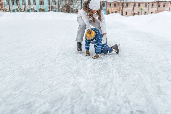 Mujer madre joven cría hijo niño 2-3 años de hielo en invierno en el parque de la ciudad. El concepto de la primera lección sobre el apoyo y el apoyo al patinaje, la ayuda de los padres en el aprendizaje de actividades deportivas y estilo de vida activo . — Foto de Stock