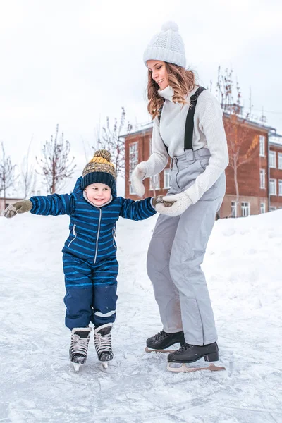 Молодая мама держит сына за руку в течение 2-3 лет, каток зимой в городском парке. Концепция первого урока поддержка и поддержка катания на коньках, помощь родителям в изучении активного образа жизни . — стоковое фото