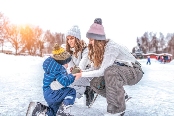 年轻的母亲、两个女人、女友抚养儿子男孩 2-3年, 在冬季城市公园溜冰场溜冰。概念第一课滑冰支持, 帮助家长学习运动和积极的生活方式. — 图库照片