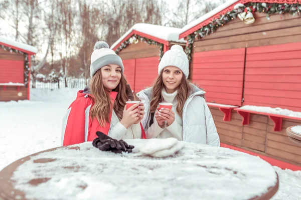 Dwie młode dziewczyny dziewczyny studentów. W zimie trzymają się filiżanki kawy i herbaty na ulicy w ich rękach. Ogrzać się w niskich temperaturach. Śniadanie i obiad w ośrodku narciarskim. Kobieta komunikować się i uśmiech. — Zdjęcie stockowe