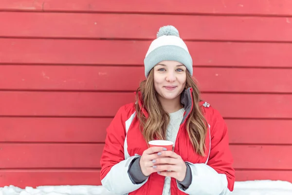 Seorang gadis cantik dengan jaket musim dingin merah berdiri di depan jendela kayu. Di tangannya memegang secangkir kopi panas dan teh, hangat. Ruang kosong untuk teks. Konsep liburan resor, wanita bahagia tersenyum . — Stok Foto