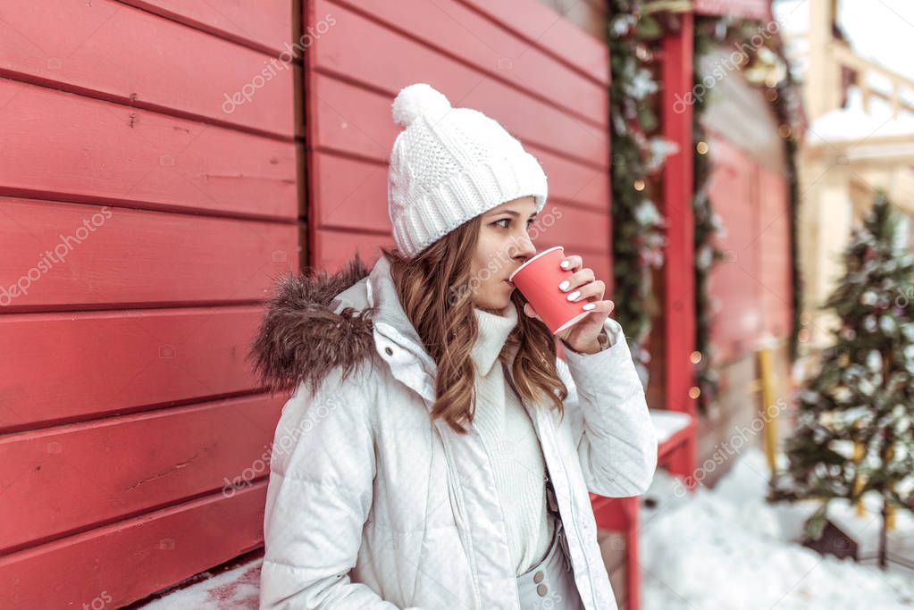 A girl in white jacket and winter hat. Standing at red wooden counter in hand holding a mug of coffee. A woman is drinking hot tea. In winter, city rests, snacks and lunch on the New Years holiday.