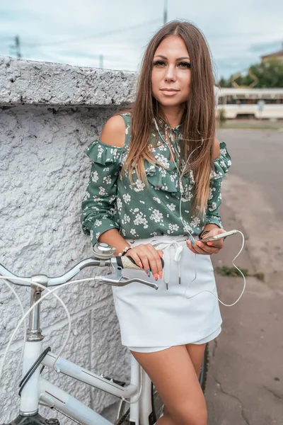 Дівчина з довгим волоссям в руці смартфон стоїть біля стіни з велосипедом влітку в місті. Прослуховування музики в навушниках . — стокове фото