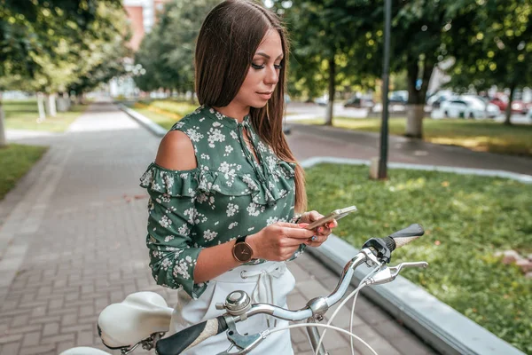 Mooi meisje met de telefoon kijken naar video's op Internet, sociale netwerken, online, Fietsen in de zomer in de stad. Een vrouw luistert naar muziek op koptelefoon. — Stockfoto