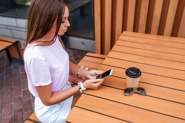 Hermosa mesa de café chica en la ciudad de verano. En manos de teléfono inteligente, taza con té de café, brotes solares. Redes sociales, en línea, correspondencia en aplicación telefónica, esperando orden en restaurante . —  Fotos de Stock
