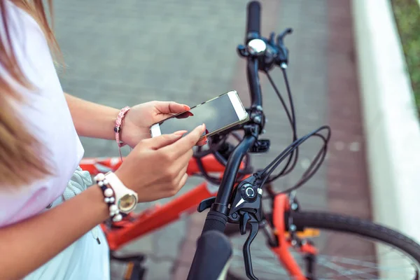 Девушка держит смартфон, ищет маршрут через приложение на карте Интернета. На фоне красного чёрного велосипеда на парковке. Онлайн в социальных сетях, коммуникационная корреспонденция . — стоковое фото