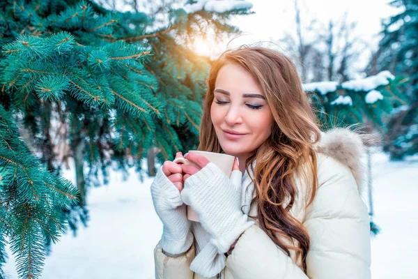 Krásná brunetka zelené pozadí zasněžené vánoční stromky drží hrnek čaje kávy bílé rukavice. Pocity štěstí potěšení oteplování horký nápoj. Koncept zimní rekreaci snídani a oběd. — Stock fotografie
