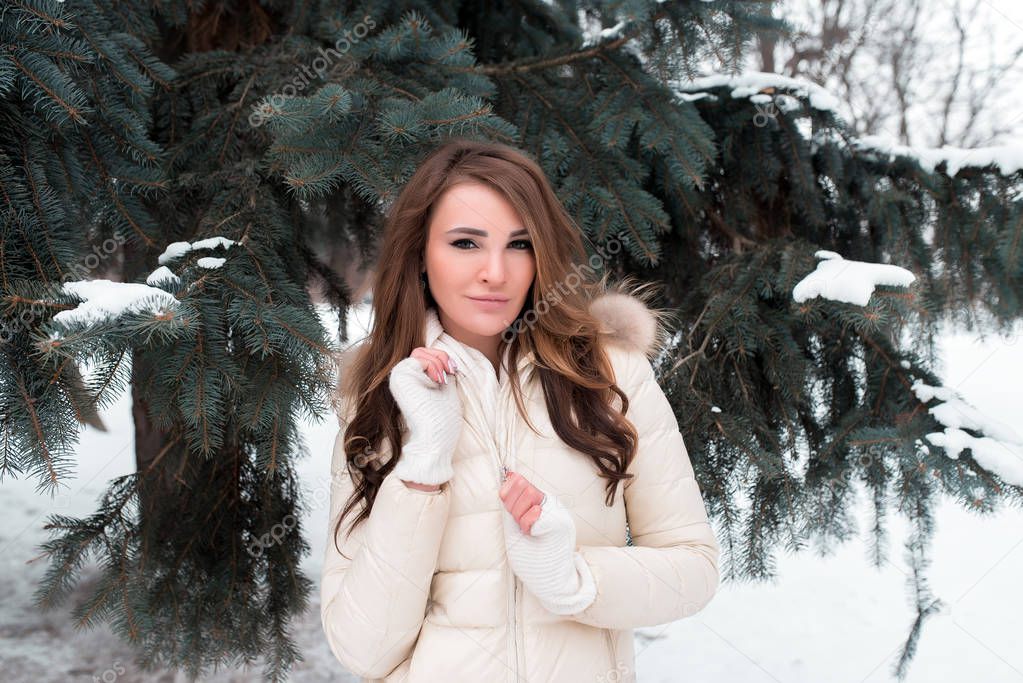 A girl in a winter park on a background of green trees, in a white down jacket with long brown hair, a happy woman smiles at the coat of lightning.