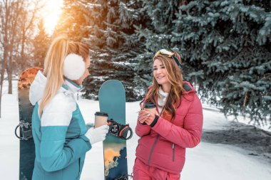 Yeşil Noel ağaçları kış park arka plan konuşurken iki kız kız snowboards, arka plan üzerinde. Kahve bardağı çay elinde. Kavramı en iyi arkadaş kız gülmek konuş. Resort adlı dinlenme.
