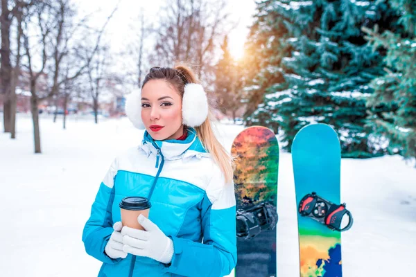 Beautiful girl in blue jumpsuit background green snow Christmas trees, snowboard boards. In hands holding mug of hot tea coffee. Emotions happiness, pleasure of rest at resort, fresh air in nature. — Stock Photo, Image