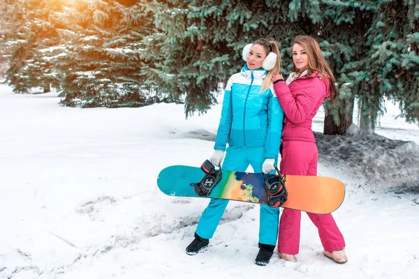 Due ragazze amiche su snowboard, in posa su sfondo di alberi di Natale verdi. Nelle mani di uno snowboard. Concetto migliori amici sorelle divertenti si rilassano sul resort. Spazio libero per il testo . — Foto Stock