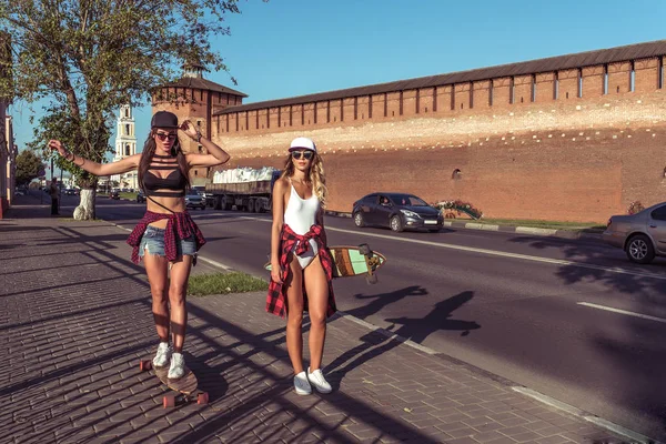 Dua gadis pacar, berjalan dan naik papan seluncur panjang, wanita bersantai untuk bersenang-senang, bahagia tersenyum santai di musim panas di kota. Terhadap latar belakang mobil jalan dan bangunan bata . — Stok Foto