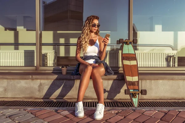 Beautiful girl with long hair writes a message in phone, online chat in social networks, the summer city. Skateboard longboard, white swimsuit and sunglasses. A cup of coffee and tea. — Stock Photo, Image