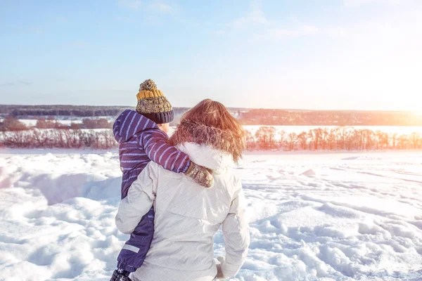 Mom with her son in her arms view from back in the winter in the city. Care and support of a small child in the open air. Against the background of snow-covered forest and the river. — Stock Photo, Image