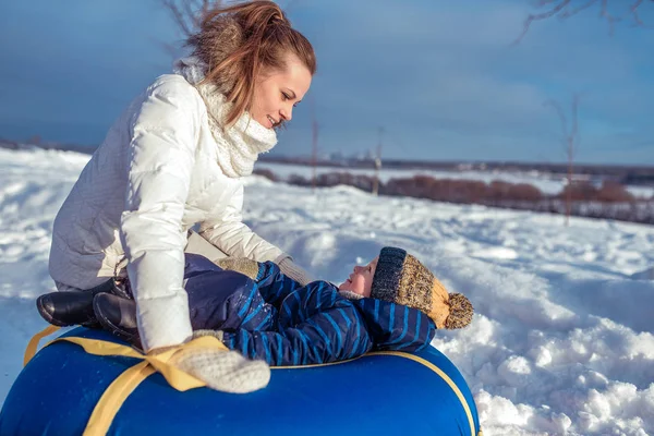 Молодая мать играет с маленьким мальчиком в течение 2-3 лет зимой на холме, сидя на синей трубке. Счастливы, глядя друг на друга. Счастливая семья играет на природе зимой в теплой одежде . — стоковое фото