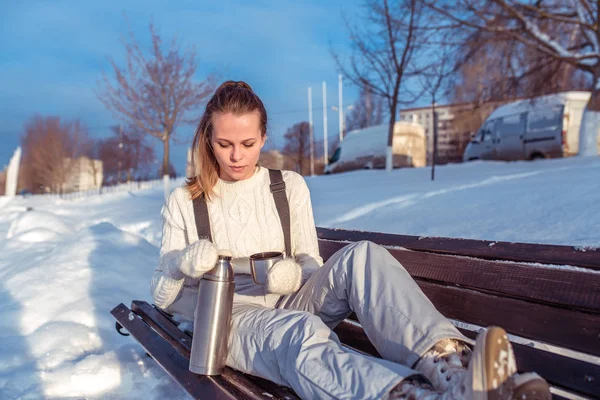 A beautiful girl pours a metal thermos with a drink, hot tea, coffee, sits on a bench in warm clothes and mittens. Holiday in the winter resort in fresh air. Warmed mug with soup. — Stock Photo, Image