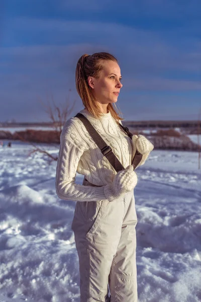 На фоне снега зимой на улице стоит красивая девушка. В белых комбинезонах в рукавицах и свитере. Смотрит на закат и встречает восход солнца. Хорошее настроение в зимний снежный день на курорте . — стоковое фото