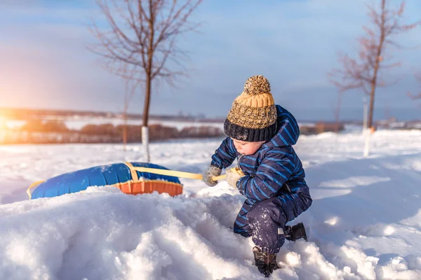 Seorang anak kecil, 2-3 tahun, menyeret mainan musim dingin, menarik dari salju tertutup snowdrift dari musim dingin taman bukit. Konsep kekuatan keberanian kepercayaan diri. Dalam topi musim dingin dan anak-anak hangat overall . — Stok Foto