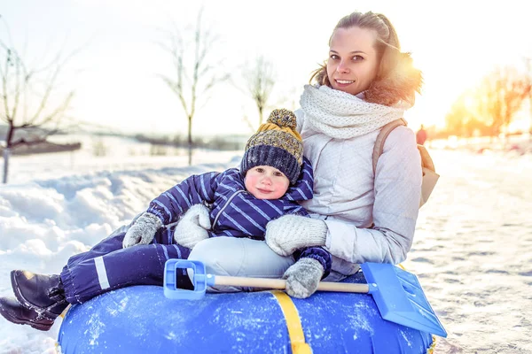 A beautiful and young mother with her son, boy 2-3 years old, sitting inflatable tubing toy. Posing camera. Rest in winter resort happy and young family is resting in warm casual clothes.