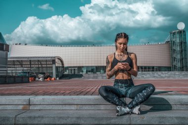 Güzel genç kız dövmeler, yaz şehir el smartphone dinlediği müzik kulaklık, spor giyim. Merdiven üstünde oturuyor. Metin için özgür yer. Internet sosyal ağlarda online. Sabah fitness açık hava.