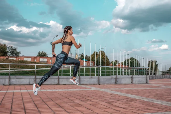 Hermosos tatuajes de niña, ciudad de verano, corre salto, en el teléfono inteligente de la mano escucha música en los auriculares, sujetador polainas ropa deportiva. Texto de espacio libre. Ejercicio matutino al aire libre sobre fondo cielo azul . — Foto de Stock