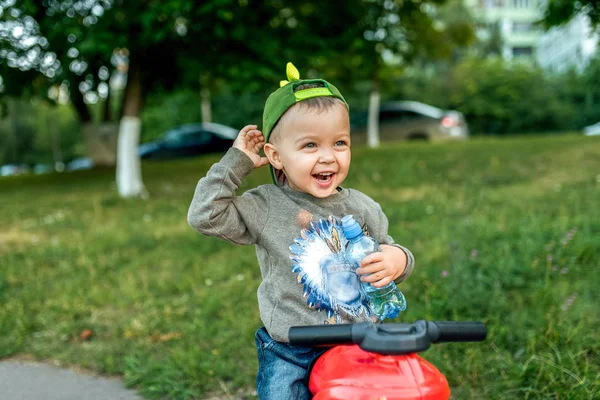 Anak laki-laki kecil berusia 3-5 tahun, duduk di atas mainan sukacita tangan ke atas, tertawa gembira di tangannya memegang botol air. Di musim panas di kota di udara segar . — Stok Foto