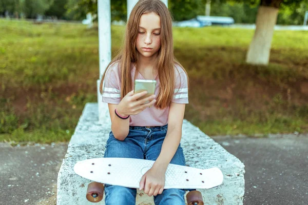 Gadis remaja 12-13 tahun, duduk di tangan skate. Di musim panas di kota dengan celana jeans dan kaos merah muda. Komunikasi di Internet, di tangan smartphone memegang menulis pesan dalam aplikasi . — Stok Foto