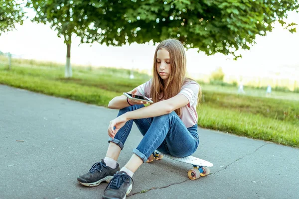 Gadis remaja berusia 11-14 tahun, duduk di tangan skate. Di kota musim panas dengan celana jeans dan kaos merah muda. Komunikasi di Internet, di tangan smartphone memegang menulis pesan dalam aplikasi . — Stok Foto