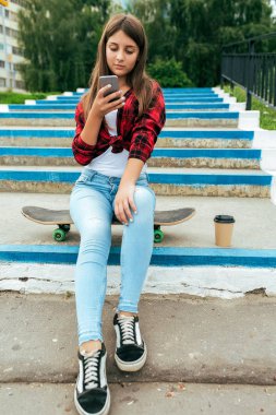 Kız kız öğrenci 10-15 yaşında, yaz şehir okul dersleri sonra dinlenme. Elinde telefon, paten oturur online internet uygulaması, sosyal ağ sohbet sohbet. Bir fincan kahve çay adımlar.