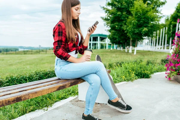 Niña colegiala de 10-15 años, ciudad de verano descansando después de las clases de la escuela. Banco en manos teléfono, aplicación de Internet en línea, correspondencia de redes sociales en el chat. Sobre el fondo de hierba y árboles . —  Fotos de Stock