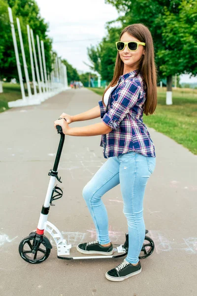 Menina adolescente estudante 10-15 anos. Em uma scooter no parque da cidade de verão. Feliz sorrindo em óculos de sol. Descansando depois da escola montando uma scooter no fim de semana. Em roupas casuais, camisa e jeans em tênis . — Fotografia de Stock