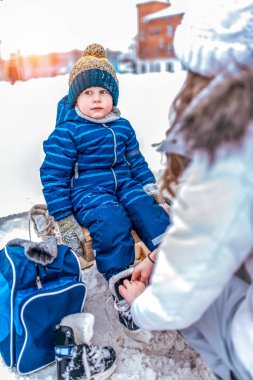 Anne oğlunu Kış Ayakkabı Kayak sonra ayakkabılar. 3-6 kentin genel bir buz pateni pisti üzerinde kışın bir bankta oturan yaşında, küçük çocuk. Mavi tulum ve sıcak bir şapka.