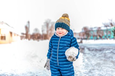 Küçük bir çocuk 3-6 yaş mavi tulum büyük kartopu elinde duruyor. Kış City snowdrifts kar ve ağaçlar arka plan üzerinde. Mutlu ayakta ve eldivenler gülümseyerek. Metin için boş alan.