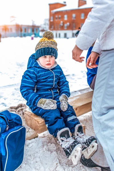 Маленький мальчик 3-5 лет, сидит на скамейке, зимой в городе на общественном катке. В синем комбинезоне и тёплой шляпе. На коньках отдыхают после катания на снегу . — стоковое фото