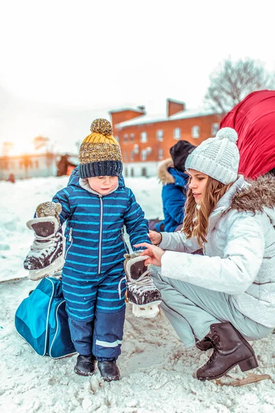 一个小男孩是 3-6, 冬天在城市的一个公共溜冰场上。穿着蓝色连身衣和一顶温暖的帽子, 手里拿着溜冰鞋。妈妈滑雪后给儿子穿冬鞋. — 图库照片