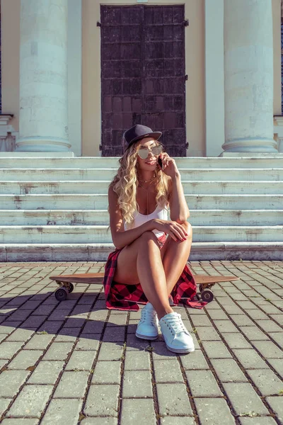 Chica bronceada en traje de baño sentada monopatín longboard. En su teléfono de mano, llamadas, hablar y sonreír, aplicación de redes sociales en línea en línea. Columnas blancas y escalones de entrada para instituir la escuela . — Foto de Stock