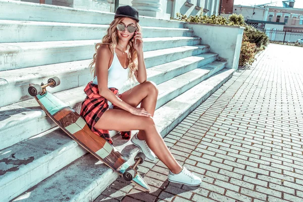 Chica bronceada en un traje de baño sentado longboard. En la mano del teléfono, las llamadas en el teléfono inteligente Internet aplicación redes sociales en línea. Pasos a la entrada del instituto o escuela . —  Fotos de Stock