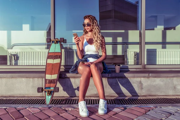 Beautiful girl sunglasses, white bodysuit background glass windows. In summer city skate, longboard. Hand phone, writes message Internet, online application social networks. Long hair cup coffee tea.