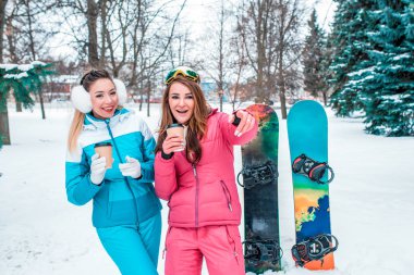 İki kız arkadaşım kız kardeşler orman kar ve snowboard gülümsüyorsun. Bir şaka ya da bir şaka komik bir şaka kendi elleriyle gösteriyorlar. Sıcak kahve veya çay Kupası elinde.