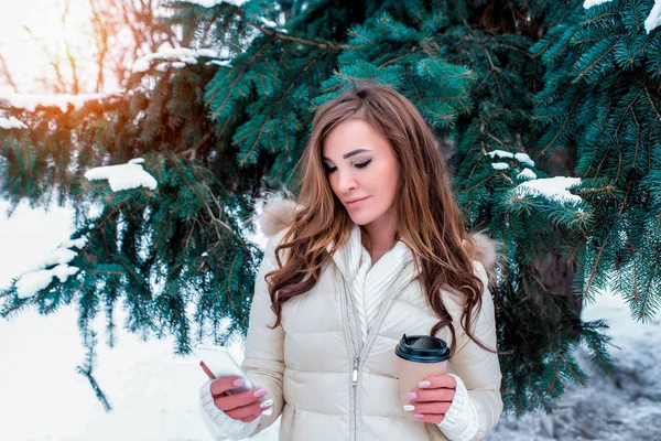 Bella ragazza nella foresta invernale con i capelli lunghi in mano che tiene una tazza, un telefono cellulare che legge il messaggio applicazione online nei social network, sullo sfondo di alberi di Natale innevati . — Foto Stock