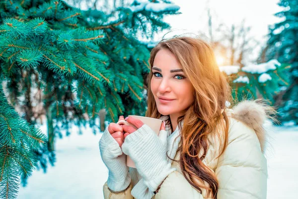 Glad och leende flicka i en vinterjacka med långt hår. I hans händer som håller en mugg hett kaffe eller te. Mot bakgrund av snöiga gröna julgranar. Vila på resort i vinter. — Stockfoto