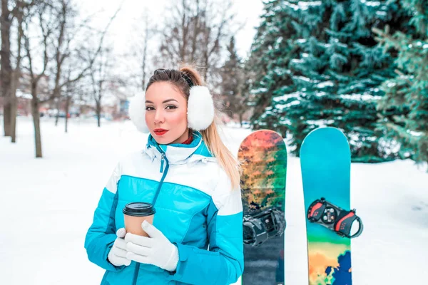 A girl with cup tea coffee, in white warm ears, is standing in a winter park against background of Christmas trees and snowboards. Rest at resort in snowy weather. Blue overalls and bright lipstick. — Stock Photo, Image