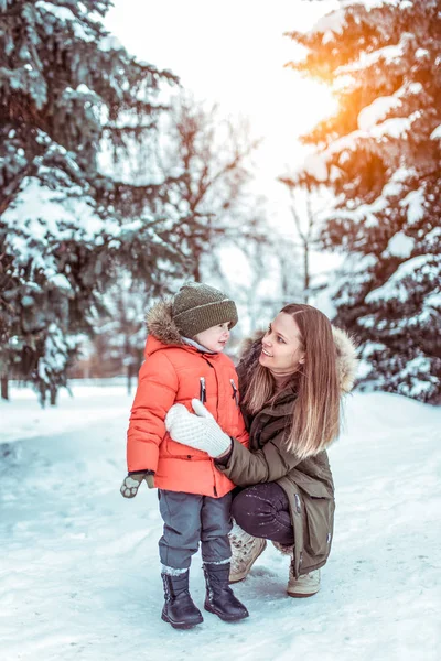 Молодая мать держит сына 3-5 лет, разговаривая зимой в лесном парке, на фоне деревьев и снежных заносов. Свободное место для текста. Радость, улыбки и веселье на зимнем курорте . — стоковое фото